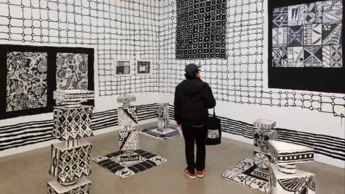 A young man wearing black clothes stares at the monochromatic totems and wall-wide floral and geometric canvases shown in a gallery.