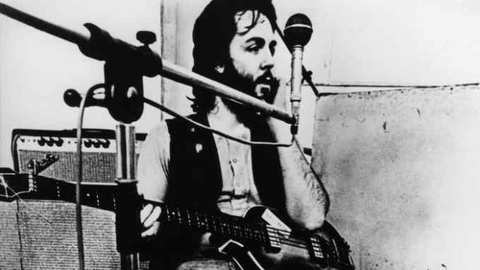 A bearded man with a bass guitar stands next to a microphone in a recording studio