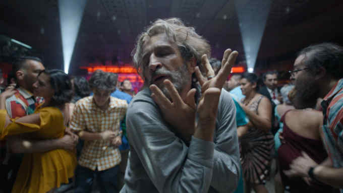 A middle-aged man with a beard and long greying hair dances expressively in a nightclub