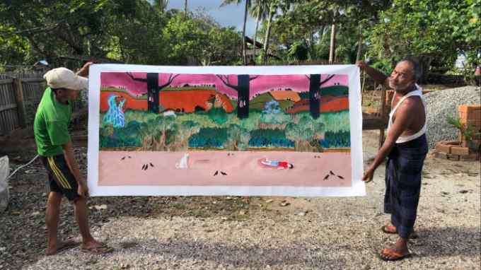 Two men stand in a yard. They hold aloft a wide acrylic painting that shows a man lying on a pink-toned beach as his white dog looks on
