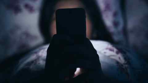 A woman lies in bed in the dark, holding a mobile phone whose illuminated screen casts a light around her