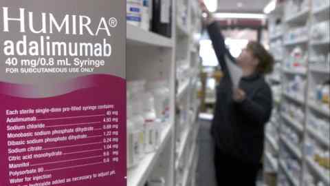 A sign for Humira in a pharmacy while a person in the background reaches up to get something off a shelf
