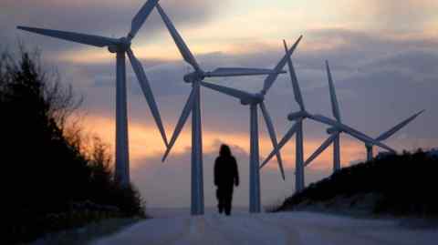 A silhouetted person walks up a road with wind turbines and a sunrise in the background