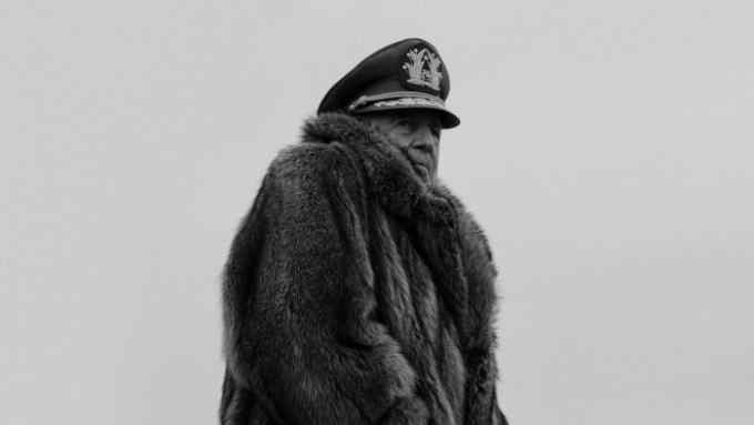 A man in a military cap and a fur coat stands huddled against the cold