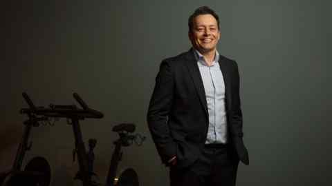 Cesar Carvalho , CEO of Gympass, wearing a coloured shirt and suit, poses in front of a static bike