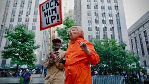 A protester outside the court wearing a costume of prison clothing, a mask of Donald Trump as another protester holds a sign that says ‘lock him up’