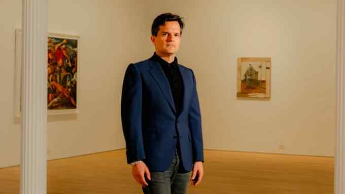 In a photo, a middle-aged man wearing a blue blazer over a black polo shirt stands before two artworks on view at a gallery.