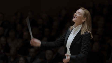 A woman in formal attire conducts an orchestra with a baton, smiling