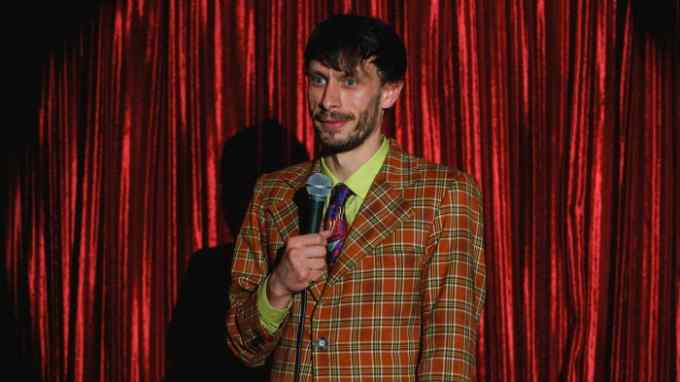 A bearded man in a checkered blazer, lime-green shirt and multi-coloured tie stands in front of a red curtain holding a microphone