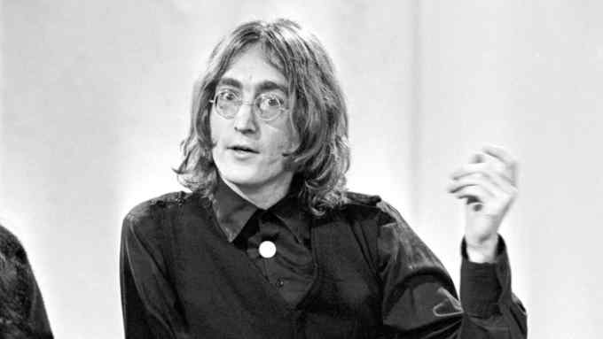 John Lennon, with long straggly hair, sits talking, gesturing with one hand
