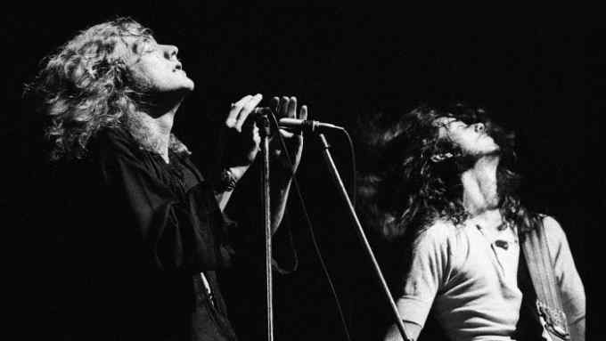 A long-haired man on stage sings into a microphone while another long-haired man plays guitar; both have their heads tilted back at the same angle