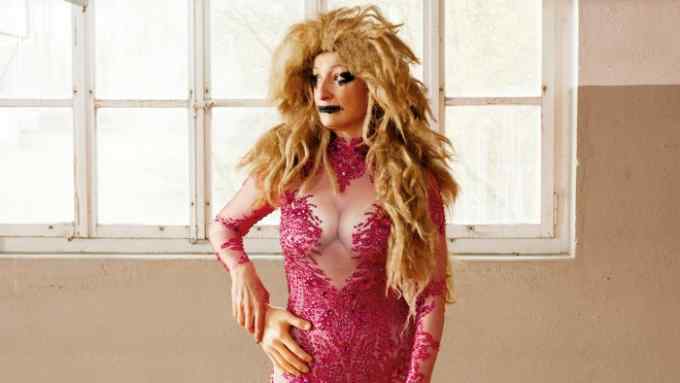 A woman posing while wearing a wig, a pink catsuit encrusted with crimson rhinestones and skates